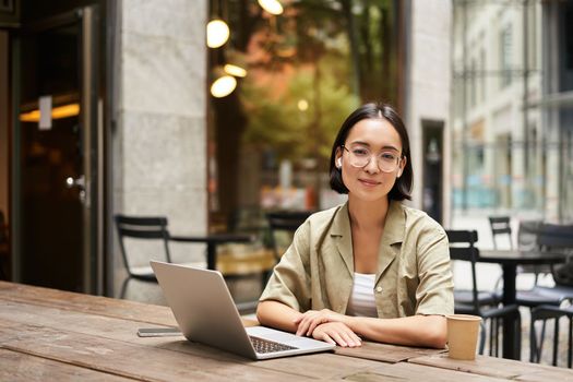 Young woman, student working from cafe, sitting with laptop and cup of coffee outdoors, looking at camera.