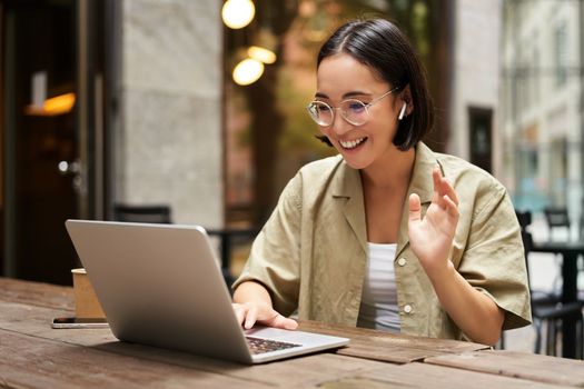 Online meeting. Young urban girl, asian woman talking with laptop, video chat, gesturing, sitting in an outdoor cafe and working remotely.