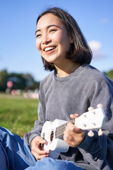 Vertical shot of smiling asian girl singing and laughing, playing ukulele, learn how to play instrument, sitting outdoors in park.