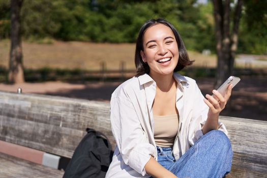 Portrait of happy young asian woman talking on mobile phone in park, sitting on bench and having a telephone call, chatting lively.