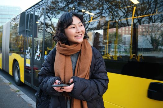 Portrait of korean girl buying ticket for public transport online, using mobile application on bus stop, wearing winter clothes.