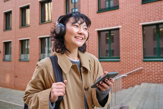 Happy young woman stands on street with backpack and smartphone, listens music in headphones, waits someone outdoors, travels around city.