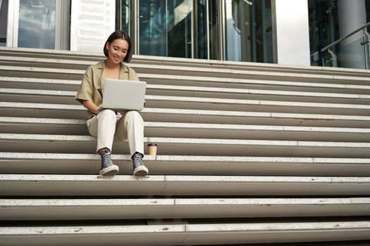 Asian girl student sits on stairs near campus, types on laptop, does her homework outdoors.