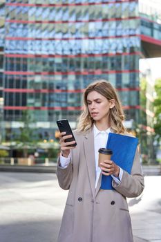 Vertical shot of corporate woman reading message on mobile phone, holding documents and drinking takeaway coffee, standing outdoors.