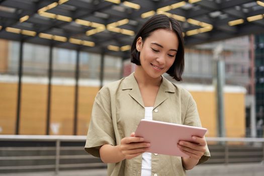 Portrait of beautiful asian girl using tablet, reading, looking at tablet while standing on street.