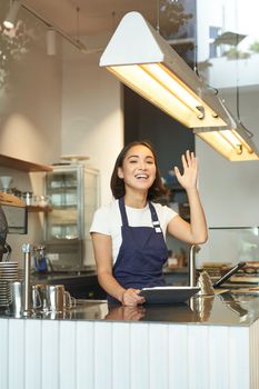 Vertical shot of happy young barista, asian cafe worker waves at client, receives orders behind counter, using tablet as POS terminal, working in coffee shop.