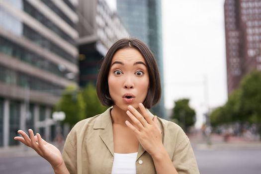 Modern asian girl standing on street with surprised face, looking impressed and amazed, hear awesome news.