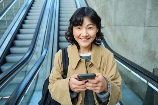 Portrait of beautiful asian girl, student goes down escalator in city, looks at mobile phone, uses telephone, map application, commutes somewhere in town.