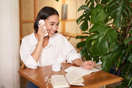 Working woman answer phone call in cafe, writing down, making notes while having conversation on telephone.