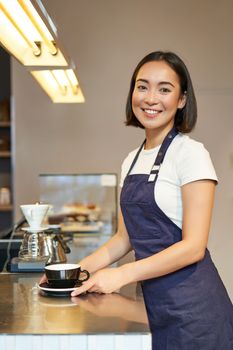 Vertical shot of smiling asian girl barista, wearing uniform, making coffee, standing near counter with cup, working in cafe.