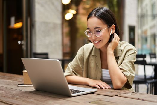 Portrait of young woman video chatting, having online meeting on laptop, sitting in outdoor cafe with compute, wearing glasses and smiling.