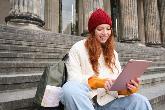 Outdoor shot of young stylish redhead girl sits on staircase and connects to public wifi, uses digital tablet, reads news on gadget.