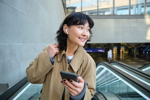 Young brunette woman commutes, goes somewhere in city, stands on escalator and uses mobile phone, holds smartphone and smiles, listens music in wireless headphones.