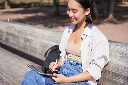 Young asian creative girl drawing with pen on graphic tablet, sitting outdoors in park, painting smth.