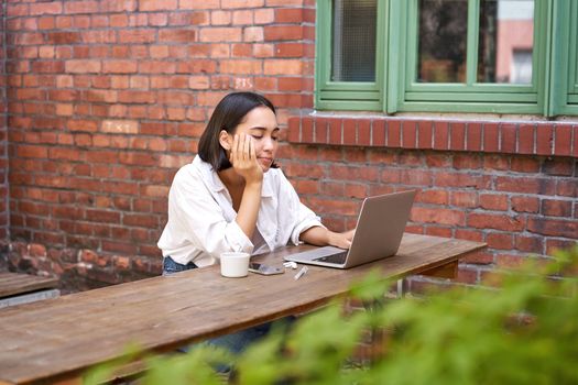 Portrait of stylish young woman, brunette girl with laptop, sitting outdoors and using computer.