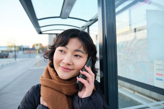 Close up portrait of cute young woman calling someone, waiting on bus stop with smartphone, using telephone while expecting public transport to arrive.