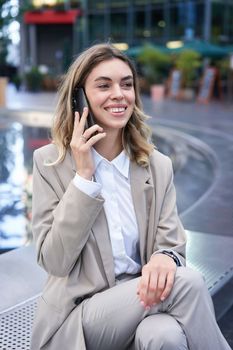 Vertical shot of smiling woman talking on mobile phone while sitting outside. Office lady waiting for someone after work.