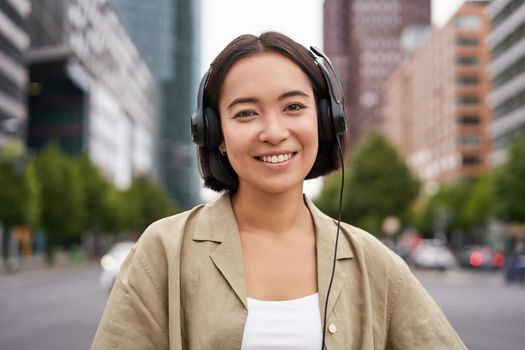 Portrait of smiling asian woman in headphones, standing in city centre on street, looking happy, listening to music.