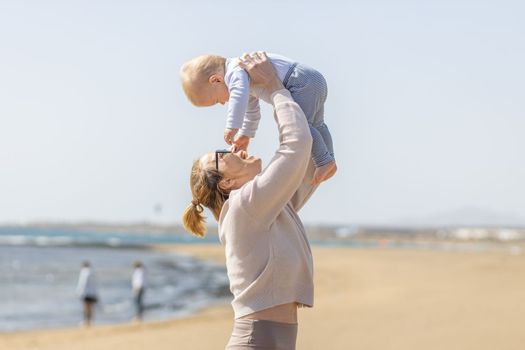 Mother enjoying summer vacations holding, playing and lifting his infant baby boy son high in the air on sandy beach on Lanzarote island, Spain. Family travel and vacations concept