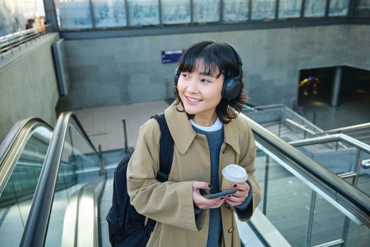 Portrait of smiling korean girl drinks coffee to go, goes up an escalator, holds smartphone, visits new city, arrives at train station, listens music in headphones.