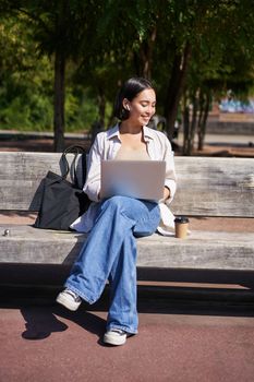 Portrait of korean woman sitting with laptop in park on bench, working outdoors, student doing homework, drinking takeaway coffee.