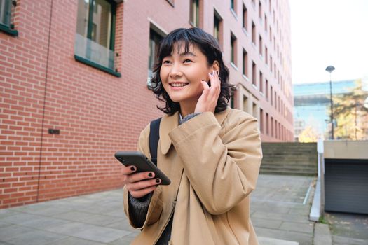 Smiling brunette woman, asian student walks on street and listens music in wireless earphones, holds smartphone, reads text message on mobile phone, picks song from playlist.
