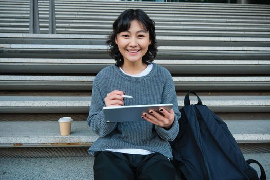 Portrait of asian girl student, hipster sitting on stairs with digital tablet and cup of coffee, draws digital art, makes design project.