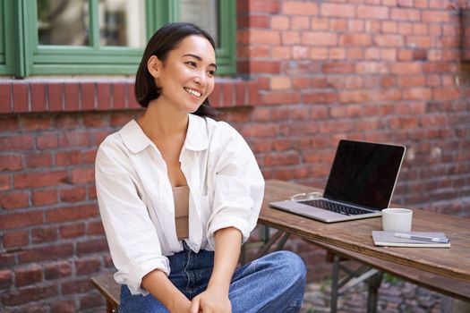 Relaxed and happy asian woman, sitting with laptop in outdoor cafe, drinking coffee, smiling and laughing.