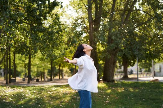 Carefree asian girl dancing, feeling happiness and joy, enjoying the sun on summer day, walking in park with green trees.