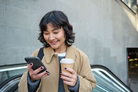 Beautiful happy asian girl, drinks coffee to go, using mobile phone while standing on escalator, walking in city centre and smiling.