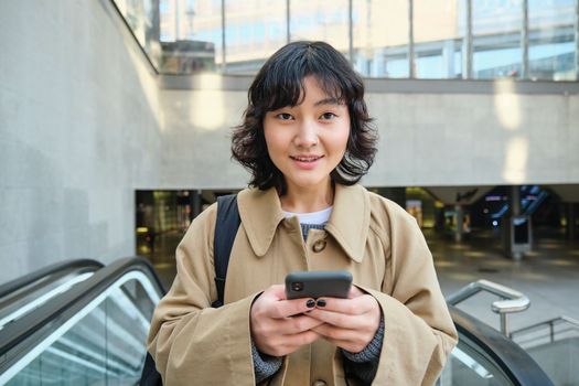 People in city. Young korean woman travels around city, goes up the escalator, uses her mobile phone and smiles.