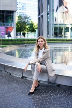 Vertical shot of successful businesswoman in suit, sitting near fountain and business hall, smiling at camera with confidence.