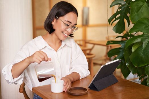 Freelance and remote workers. Smiling young woman pouring coffee in a cup, sitting in cafe and looking at digital tablet.