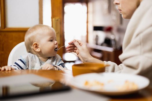 Mother spoon feeding her infant baby boy child sitting in high chair at the dining table in kitchen at home.