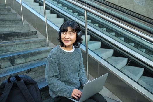 Portrait of beautiful young korean woman, student sits on stairs in public place, listens music in headphones, works on project on laptop, works remotely.