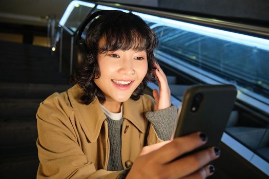 Portrait of happy asian girl in headphones, smiling and looking at smartphone, watching video or listening music on mobile phone, sitting on staircase.