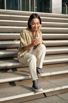 Vertical shot of asian woman, student sits on stairs in city, looking at mobile phone screen and smiling, using smartphone app.