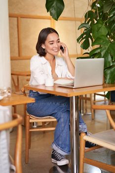 Young female cafe manager, owner sitting with laptop, answering phone calls and smiling friendly.
