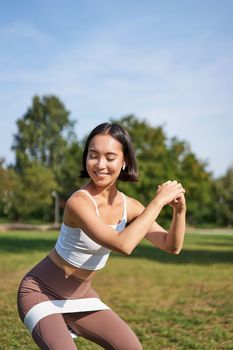 Vertical shot of young fit woman does squats in park, using stretching band on legs, smiling pleased while workout.