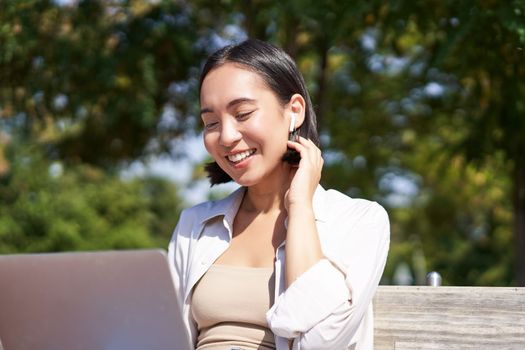 Beautiful young korean woman in wireless earphones, video chat, working outdoors in park with laptop, smiling and looking happy.