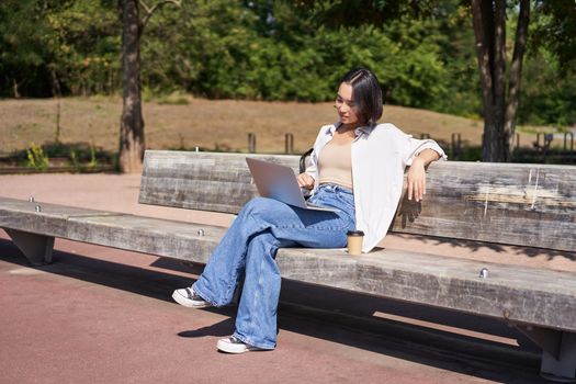 Portrait of beautiful young woman sitting on bench in park, using laptop and drinking coffee, relaxing outdoors on summer day.