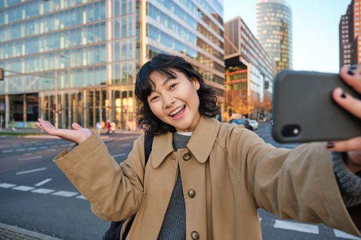 Upbeat asian girl takes selfie with smartphone in city centre, showing something behind her with smiling face. Tourist goes sightseeing.