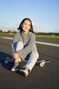 Vertical shot of asian girl skater, sits on her skateboard and smiles, enjoys sunny day, cruising on longboard on empty road outdoors.