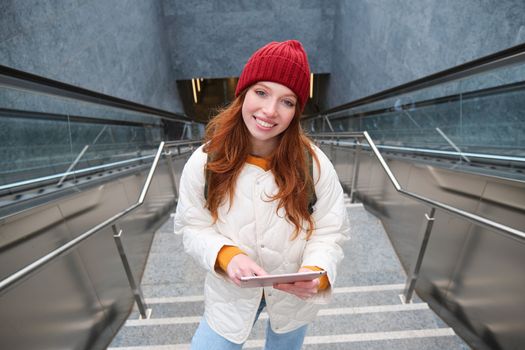 Beautiful redhead female model posing in city, walking up stairs with digital tablet, using gadget to plan her route, reading while going somewere.