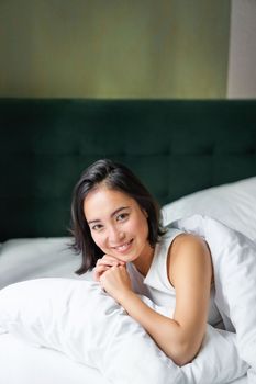 Vertical shot of smiling korean woman lying on pillow in her bed, spending morning in bedroom, waking up from sleep, looking happy.