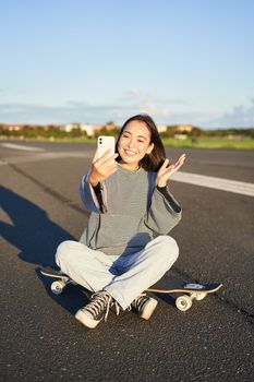 Happy teen asian girl takes selfie, video chats on smartphone app while sits on skateboard, skater records vlog while sits on longboard.