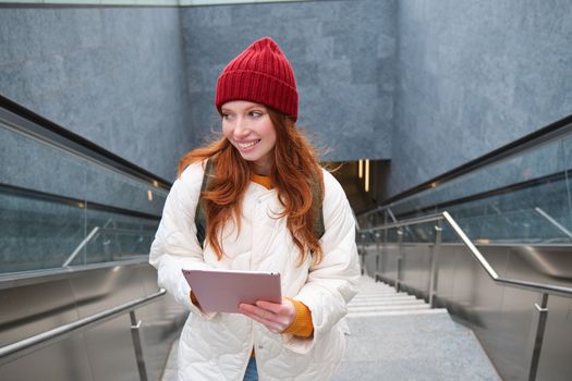 Beautiful redhead female model posing in city, walking up stairs with digital tablet, using gadget to plan her route, reading while going somewere.