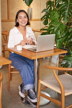 Vertical shot of young asian girl sits in cafe with laptop and smartphone, relaxing and surfing the net, working remotely.