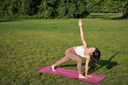 Portrait of young asian woman stretching, doing yoga on rubber mat, exercising in park, mindful training on fresh air.