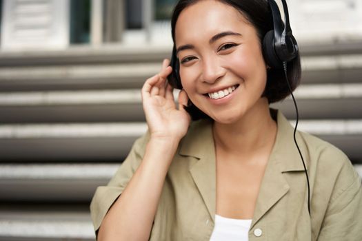 Happy asian girl listens to music in headphones, smiles, sits outdoors on stairs.